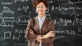 Confident female teacher standing with arms crossed in front of a detailed mathematical blackboard.
