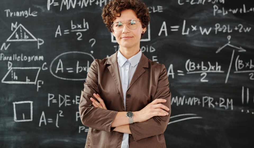 Confident female teacher standing with arms crossed in front of a detailed mathematical blackboard.