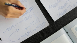 Close-up of a student solving mathematical equations with papers and textbook on a table.