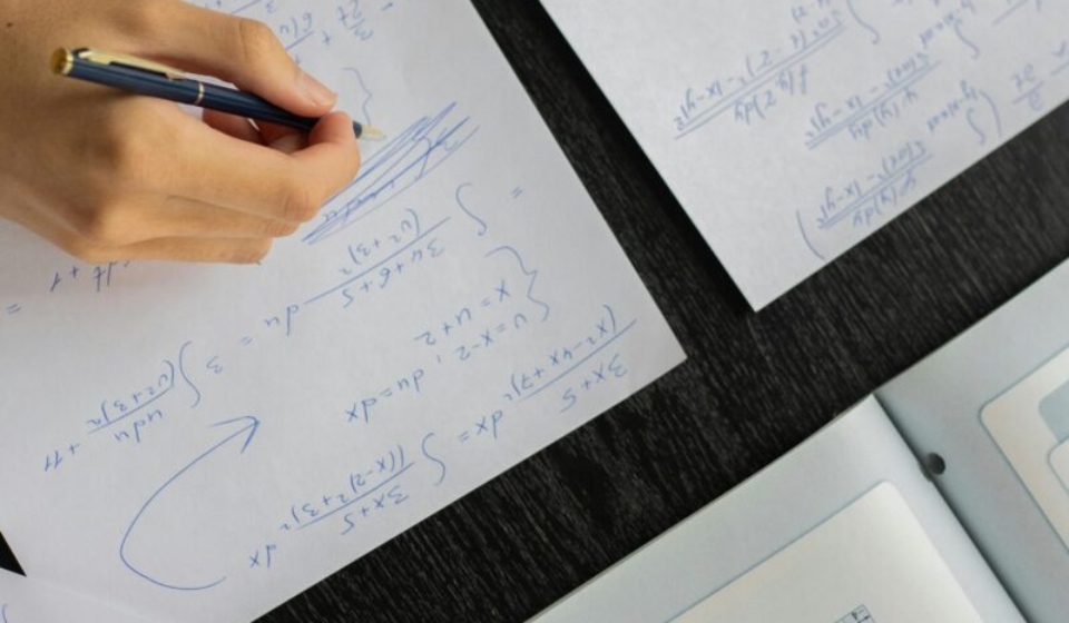 Close-up of a student solving mathematical equations with papers and textbook on a table.
